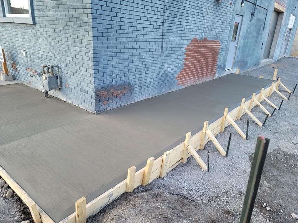 Fresh concrete walkway with timber forms running beside a blue-painted brick building