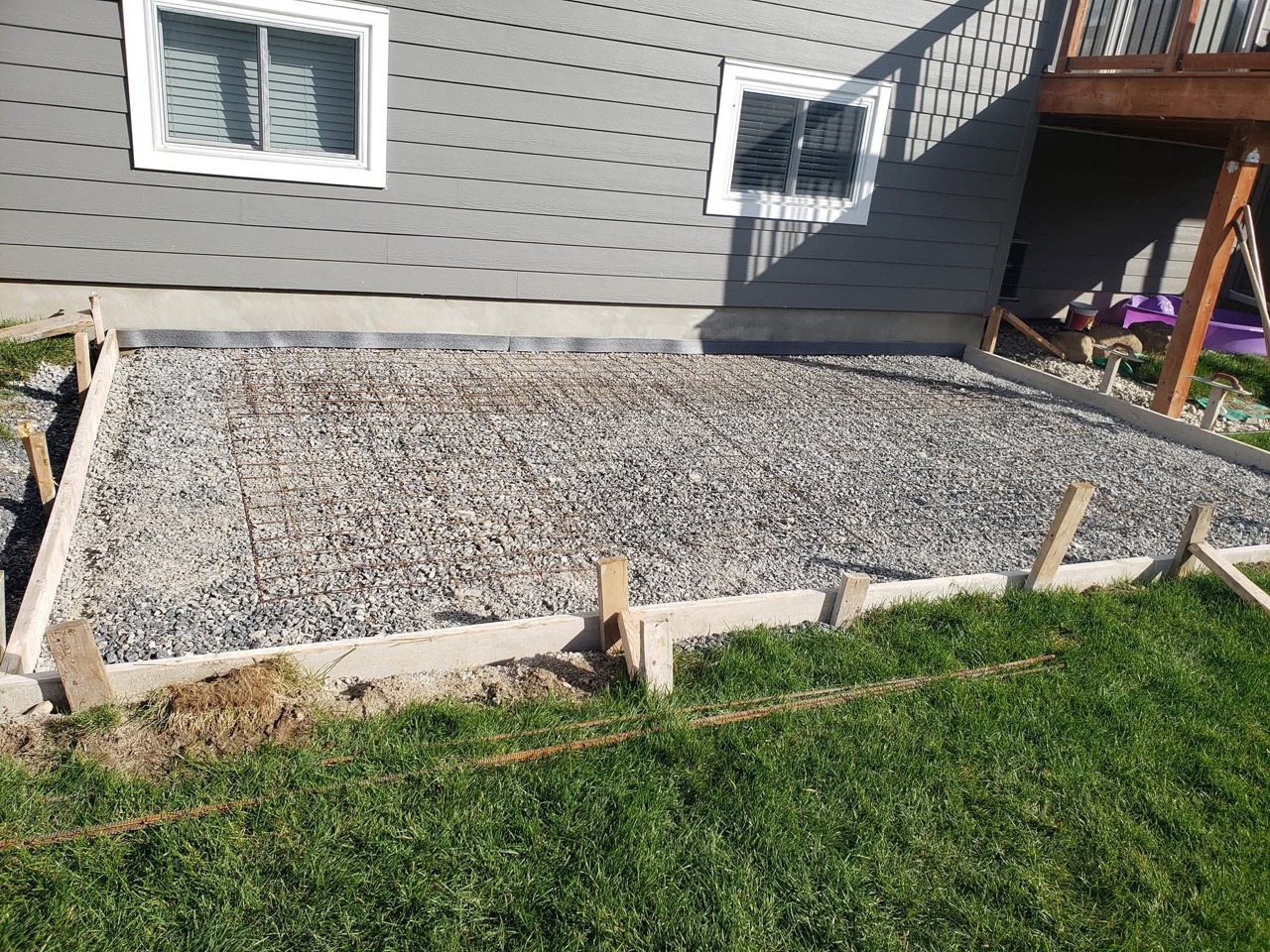 Prepared gravel base and reinforcement inside wooden forms beside a grey-sided house